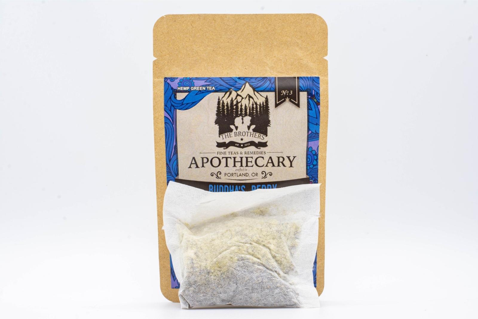 A tea bag in front of a small packet of The Brothers Apothecary's Buddha's Berry Tea on a white background