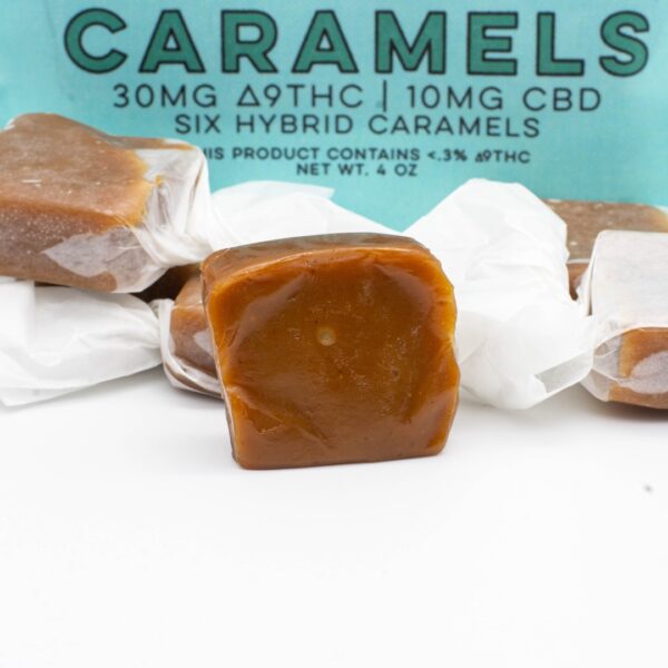 A 6 pack, next to a pile of Seventh Hill CBD's 3:1 Revive Caramel, with one caramel opened, on a white background
