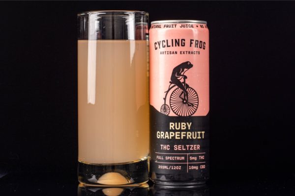 A glass of Ruby Grapefruit THC Seltzer by Cycling Frog Artisan Extracts, poured out to show contents, next to a single can, on a black background