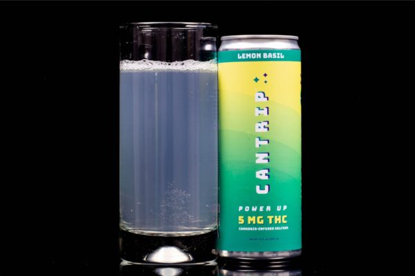A single can of lemon basil flavored hemp infused seltzer by Cantrip, next to a clear glass container displaying the drink, on a black background