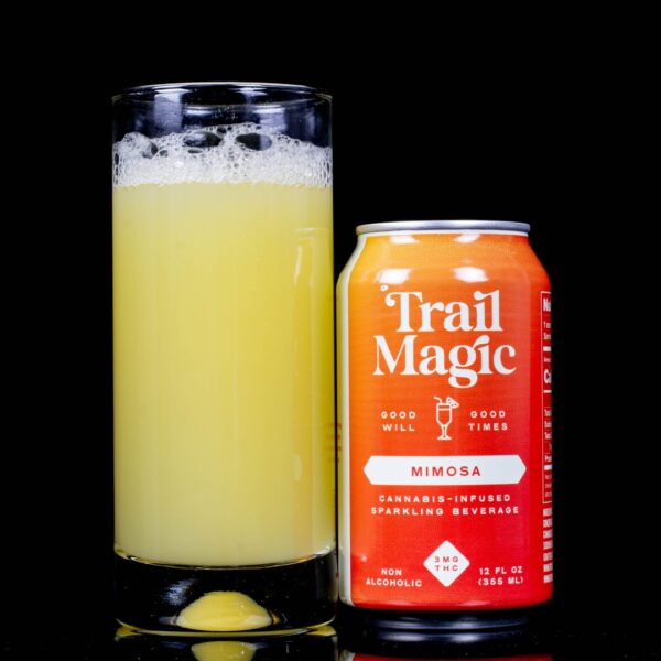 A single can of Mimosa infused sparkling beverage, by Trail Magic, next to a clear glass container containing the drink, on a black background