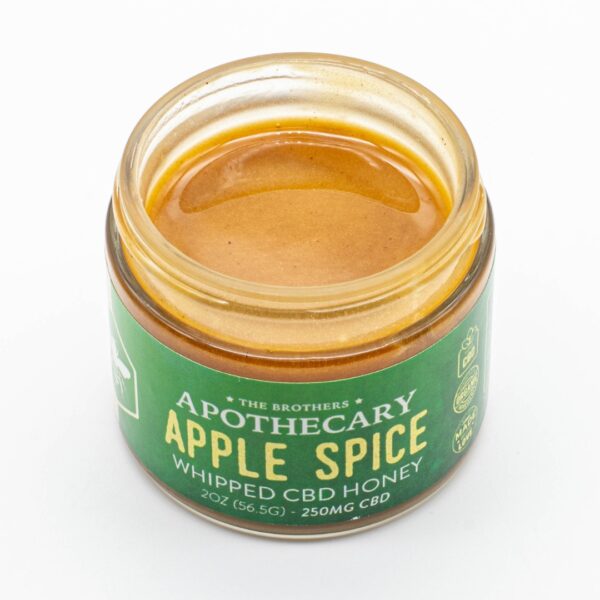 A jar of Apple Spice - CBD Honey by The Brothers Apothecary, with its lid off showing the honey, on a white background
