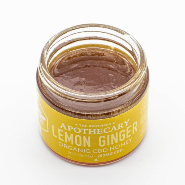 A jar of Lemon Ginger - CBD Honey by The Brothers Apothecary, with its lid off showing the honey, on a white background